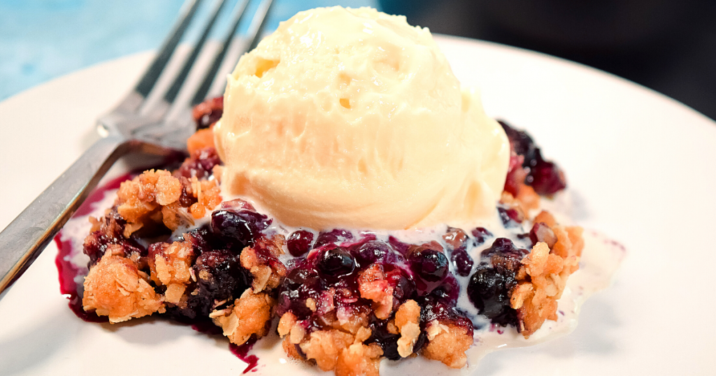 A close up image of a scoop of blueberry crisp topped with vanilla ice cream on a white plate. 