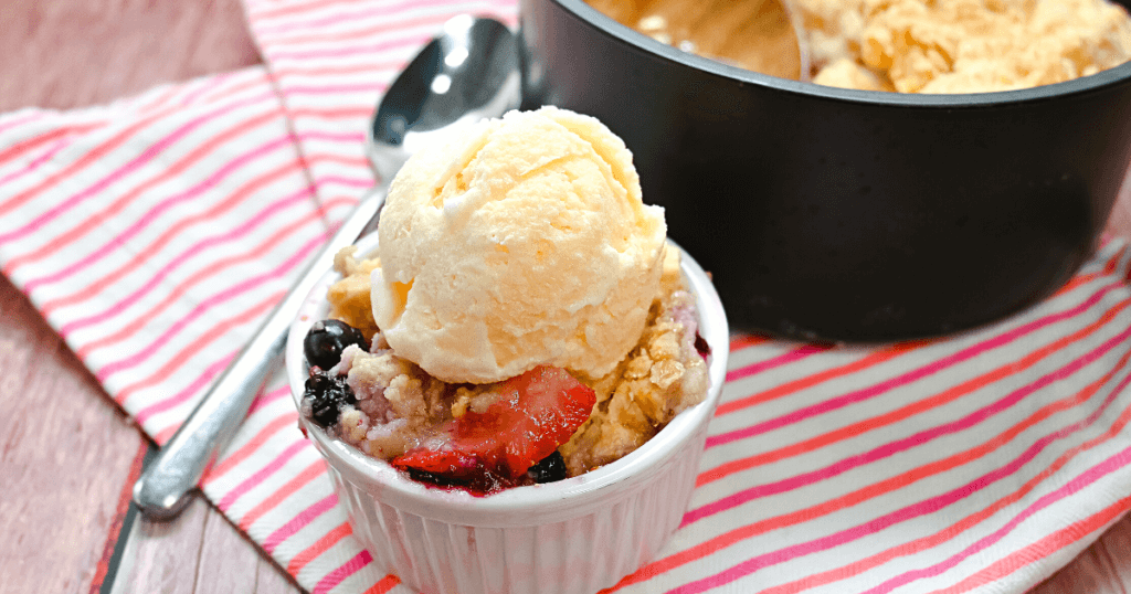 A scoop of this delicious air fryer berry cobbler in a white ramekin and topped with vanilla ice cream on a pink striped towel in front of the cake barrel with a spoon next to it. 