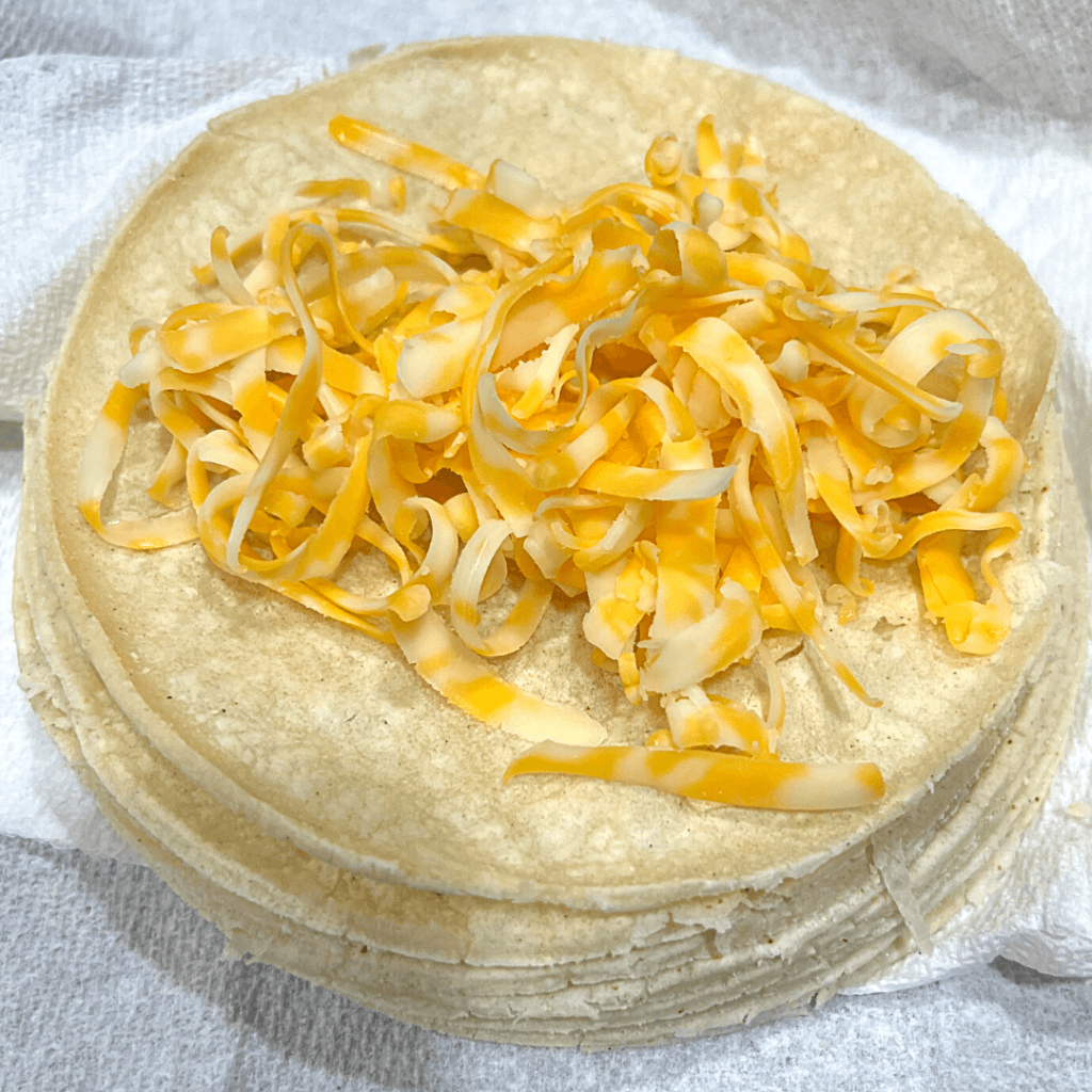 A stack of corn tortillas with the top one covered in shredded cheese and ready to roll up for enchiladas