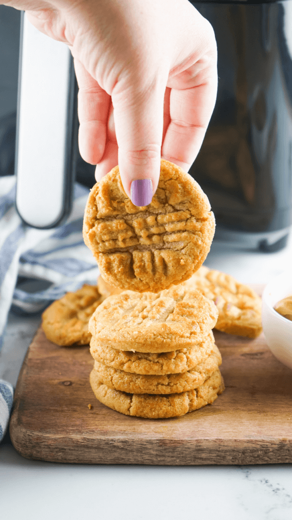 A hand with a purple fingernail holding up a peanut butter cookie on top of a wooden cutting board in front of a black air fryer. 